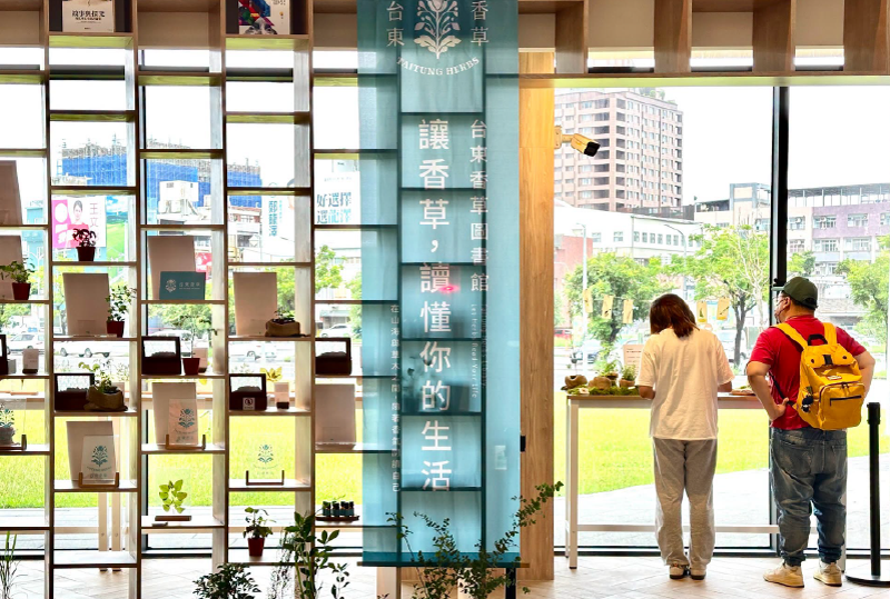 Visitors browsing the Taitung Herbs library corner beside a teal Taitung Herbs banner and potted plants